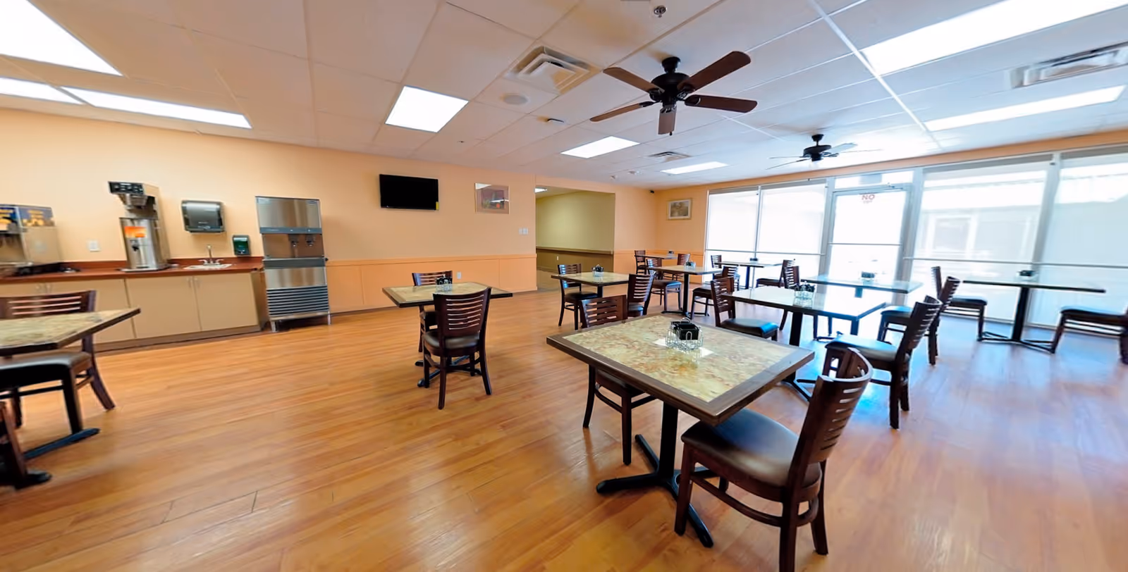 A spacious dining room with several tables and chairs arranged neatly on a wooden floor. The room has large windows allowing natural light to fill the space, ceiling fans, and a beverage station along one wall with a water dispenser and coffee machine.