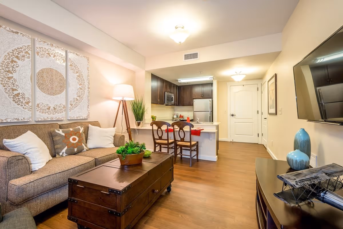 A cozy living room and kitchen area in an apartment at TerraBella Sugarloaf. The living room features a beige sofa with decorative pillows, a wooden coffee table with a plant centerpiece, and a floor lamp. The kitchen has dark wood cabinets, stainless steel appliances, a countertop with two wooden chairs, and a door leading to another room. A flat-screen TV is mounted on the wall opposite the sofa, with decorative vases and a model airplane on the TV stand.