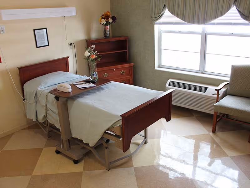 A neatly made hospital-style bed with a light blue blanket and a wooden headboard and footboard in a room. There is a small wooden dresser with flowers in a vase on top, a chair with wooden arms, a window with blinds and a valance, and an air conditioning unit beneath the window. The floor has a checkered pattern of beige and light brown tiles.