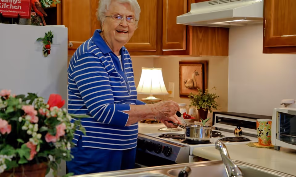 An elderly woman wearing a blue and white striped shirt is cooking in a kitchen. She is stirring a pot on the stove and smiling at the camera. The kitchen has wooden cabinets, a white stove, a microwave, a colorful mug, a lamp, and some plants and flowers on the counter.