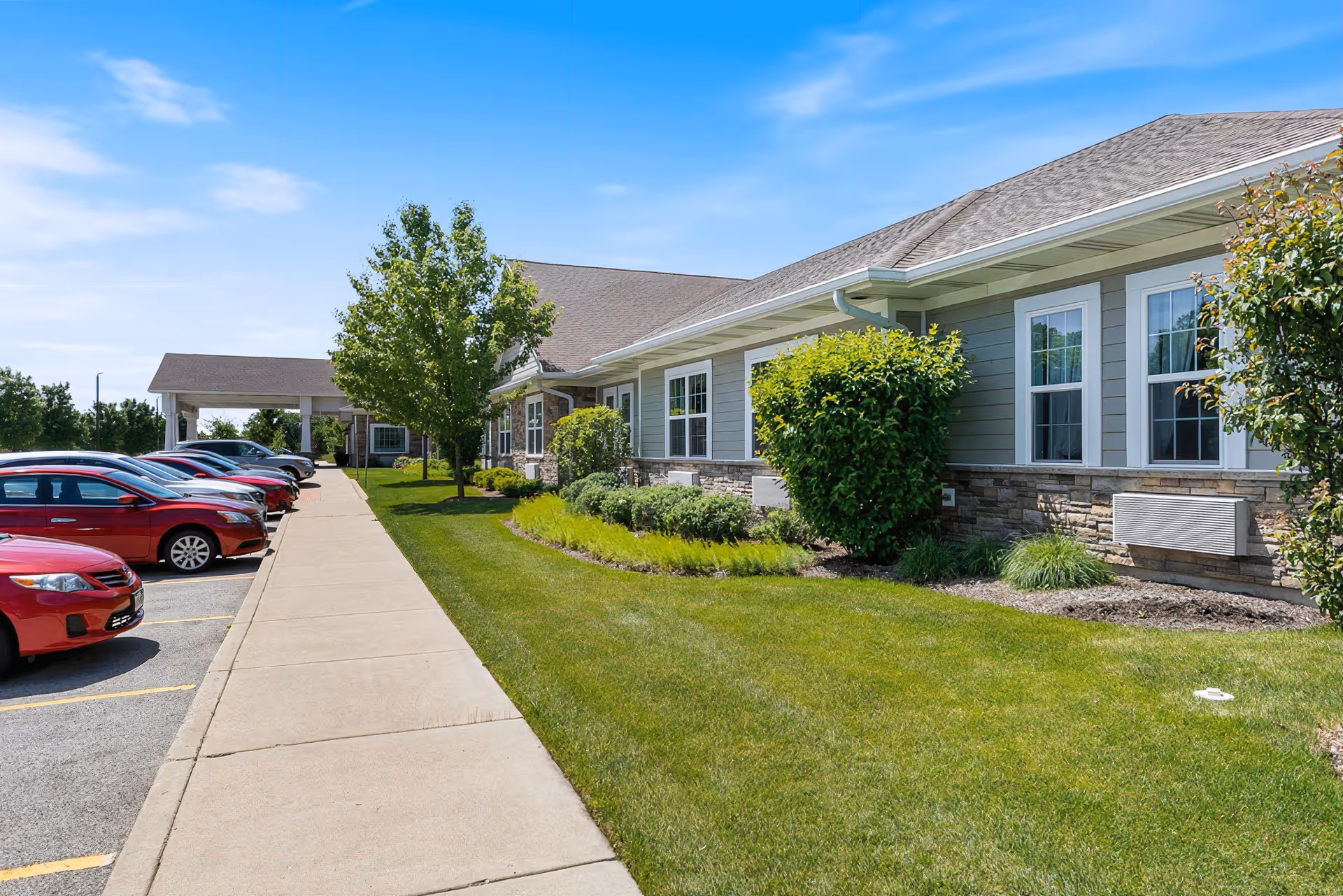 Exterior front of a single-story senior living facility with a sidewalk, landscaped lawn, parked cars, and a covered entrance.