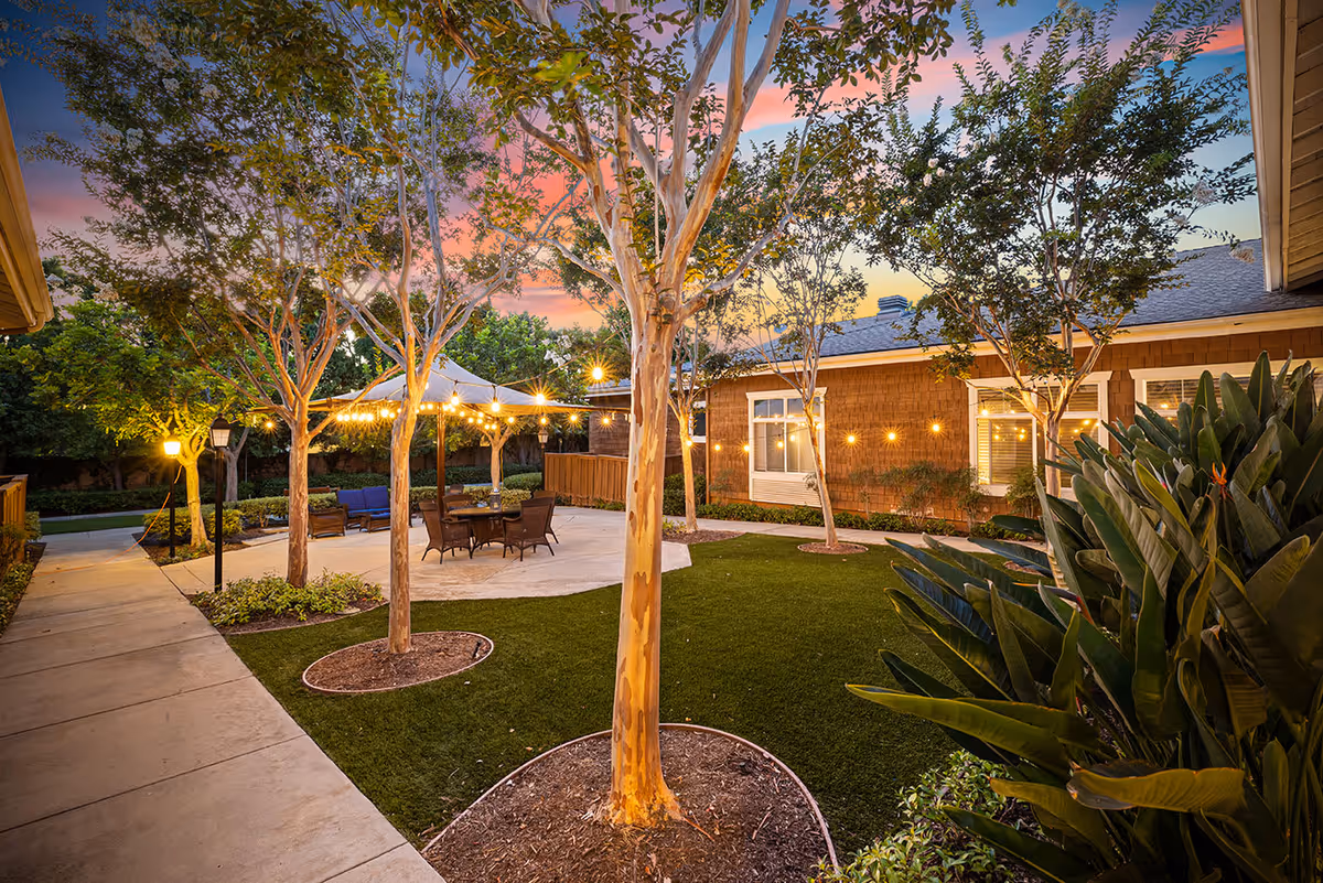 Outdoor courtyard area at sunset with trees, string lights, a paved seating area with a table and chairs under a canopy, surrounded by greenery and buildings.