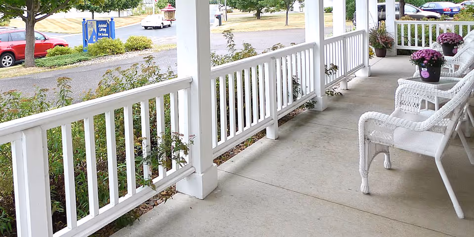 Covered front porch with white railing and wicker chairs adorned with potted purple flowers, overlooking a street.