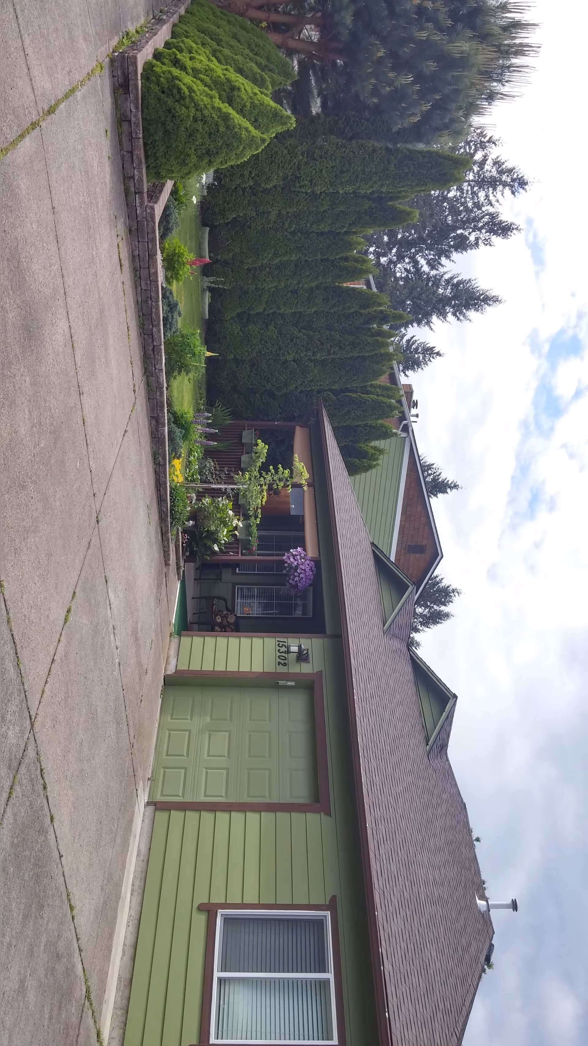 Exterior view of a green single-story house with a brown roof, a garage door, and a front porch decorated with plants and flowers. Tall evergreen trees and a well-maintained garden are visible along the side of the house. The sky is partly cloudy.