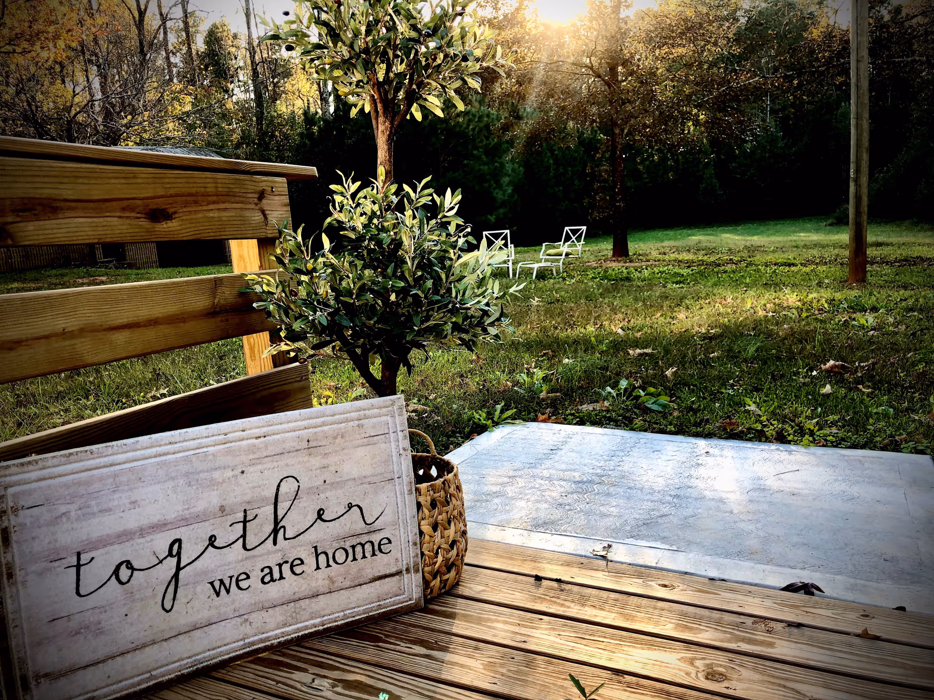 A wooden deck with a small potted plant and a decorative sign that reads 'together we are home'. In the background, there is a grassy yard with two white chairs and trees under sunlight.