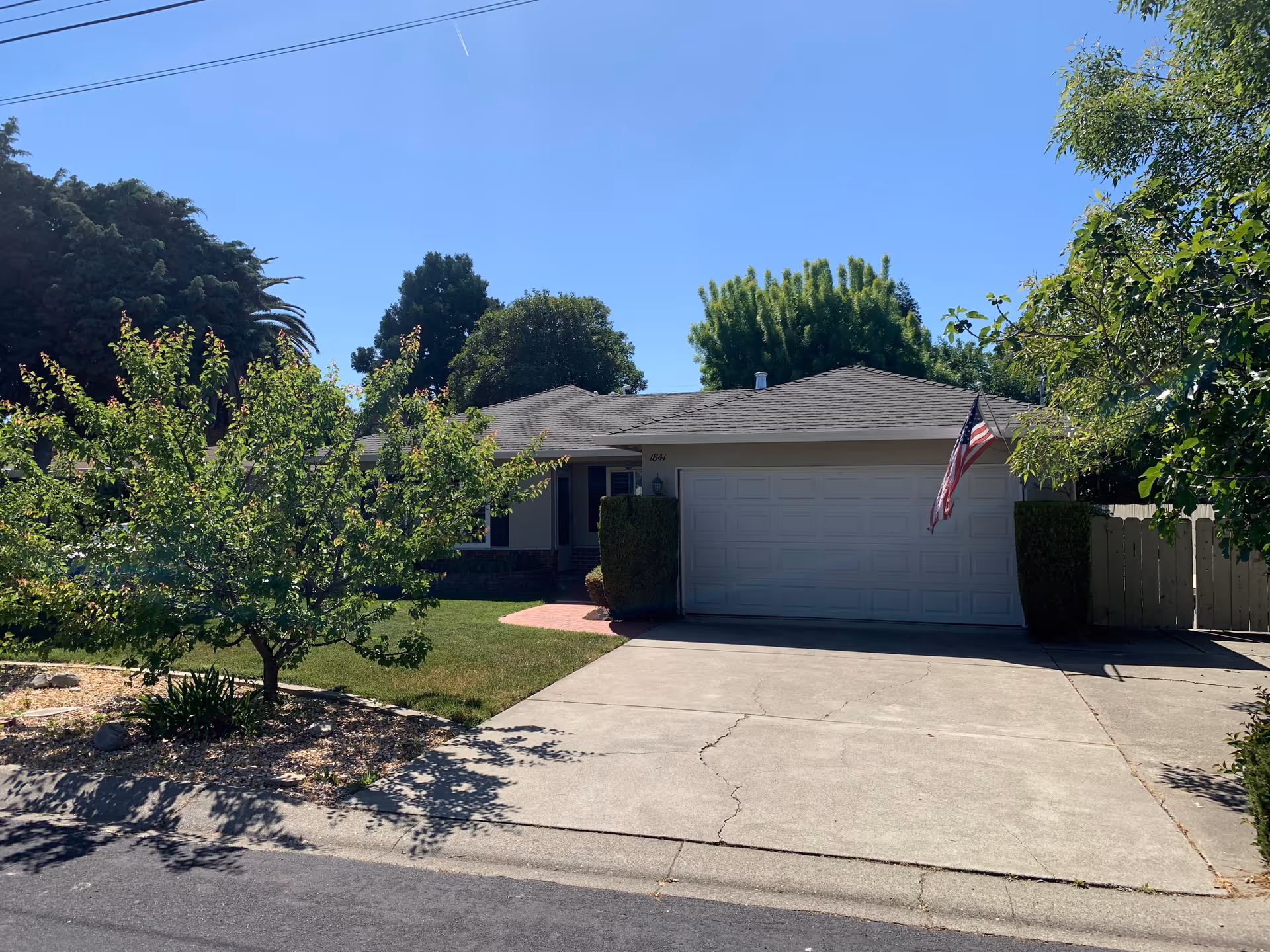 Single-story residential house with a two-car garage, an American flag mounted near the garage door, a concrete driveway, and a front yard with green grass and trees under a clear blue sky.
