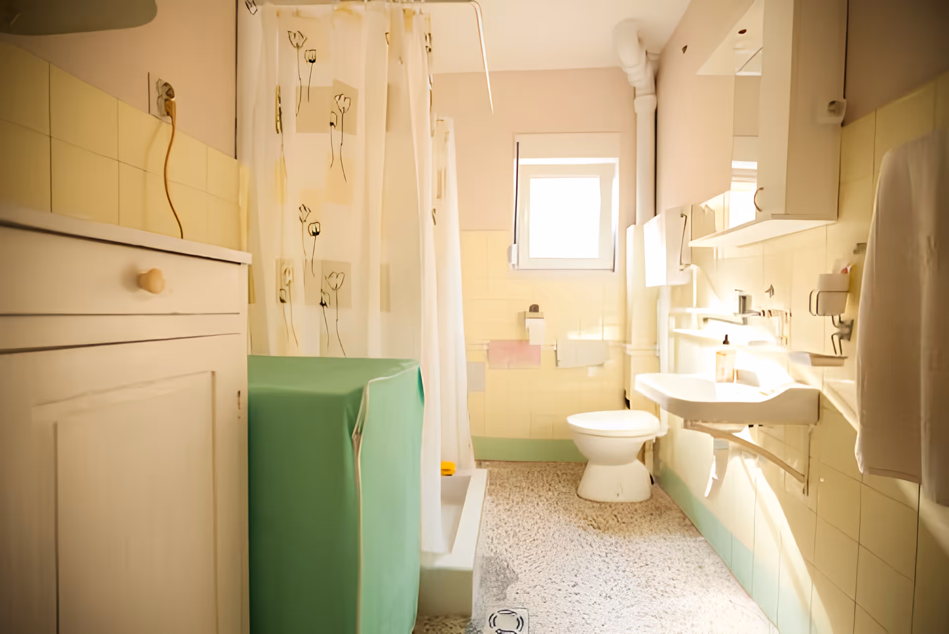 A bright bathroom with yellow tiled walls, a white toilet, a white sink with a mirror cabinet above it, a shower area with a floral-patterned curtain, and a green-covered washing machine or storage unit. There is a window letting in natural light and a towel hanging on the wall.