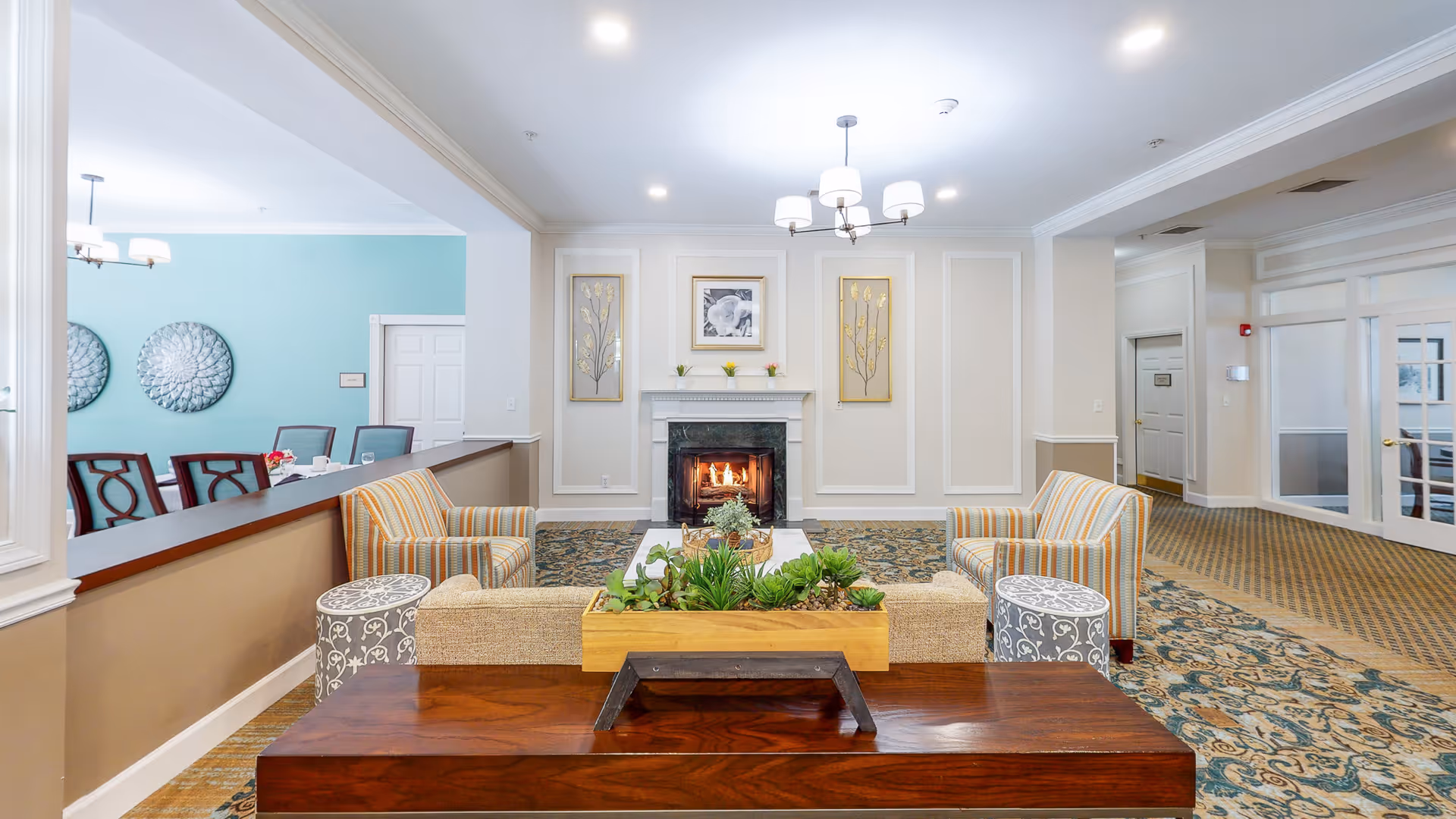 A cozy senior living facility common area with a lit fireplace centered on the far wall, flanked by two framed floral artworks. The room features a patterned carpet, two striped armchairs, a beige sofa with a wooden table behind it holding a planter with green succulents, and two small round side tables with decorative patterns. To the left, a dining area with a turquoise accent wall and dark wooden chairs is partially visible. The ceiling has recessed lighting and a chandelier with white lampshades.