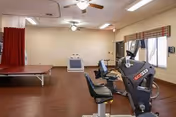 Bright rehabilitation/exercise room with two recumbent exercise bikes in the foreground, a treadmill, a treatment table behind a red privacy curtain, and a TV.