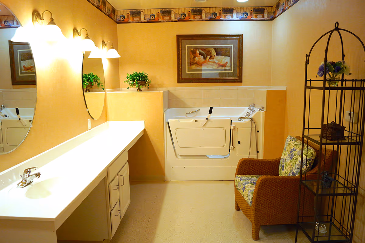 A well-lit bathroom with a long white countertop and sink on the left side, two oval mirrors above the countertop, and three wall-mounted light fixtures. At the back, there is a walk-in bathtub with safety handles. On the right side, there is a cushioned wicker chair with floral upholstery and a black metal shelving unit holding decorative items and flowers. The walls are painted a warm beige color with a decorative border near the ceiling and a framed painting above the bathtub.