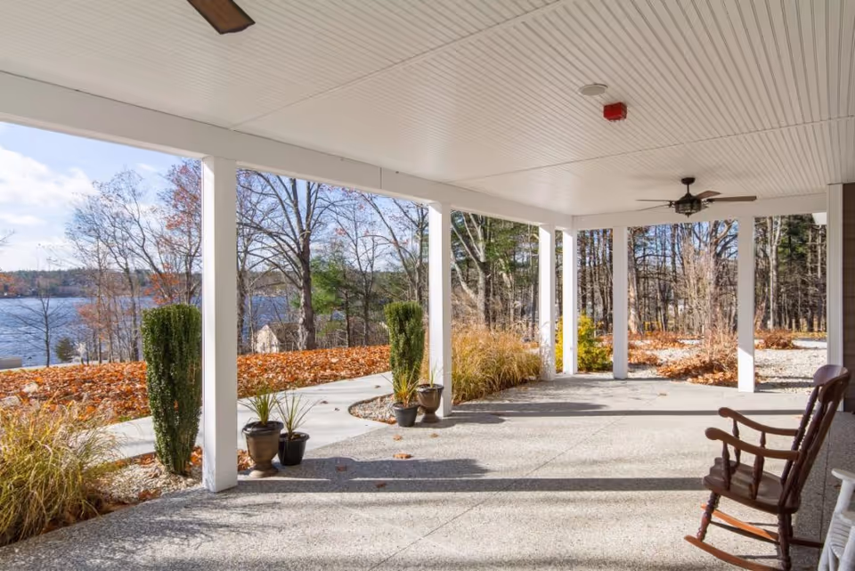 Covered outdoor patio area with white ceiling and columns, ceiling fan, potted plants, and a wooden rocking chair. The patio overlooks a scenic view of a lake and trees with autumn foliage.