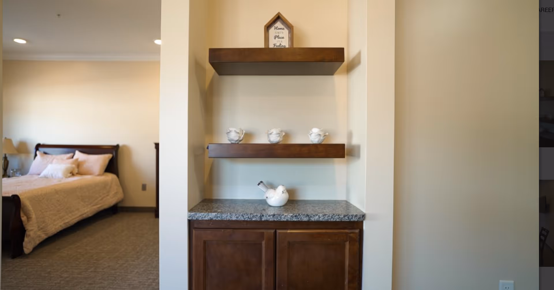 Built-in countertop and floating shelves with decorative teapots set beside a bedroom with a bed and pillows.