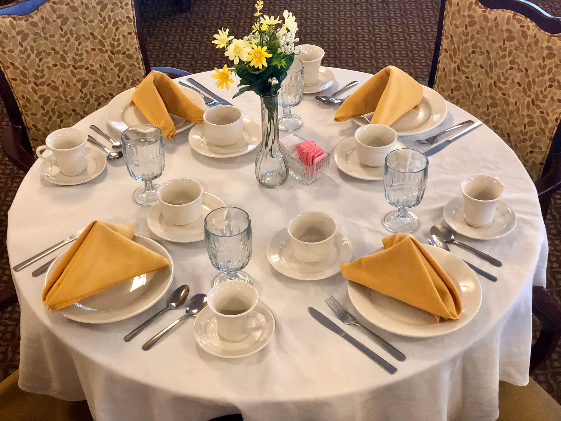 A round dining table set for six with white tablecloth, yellow folded napkins on white plates, white cups and saucers, clear water glasses, silverware, and a small vase with yellow and white flowers in the center.