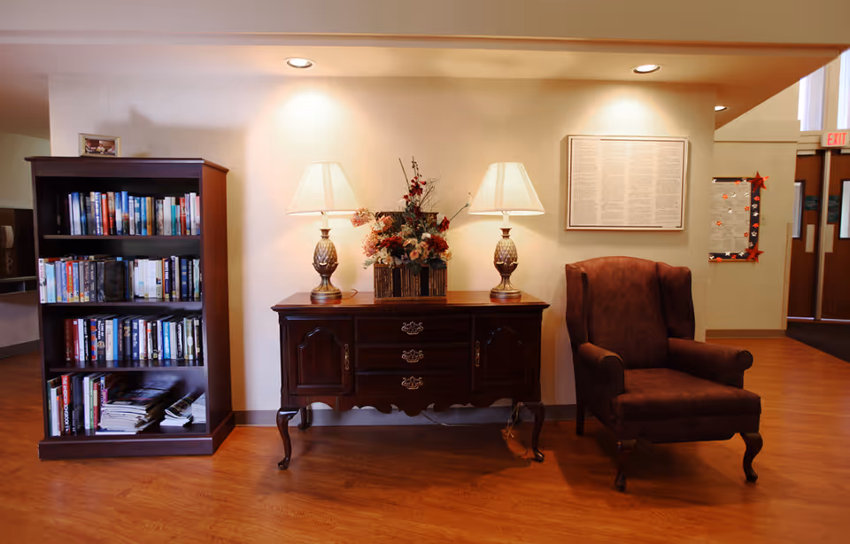 A cozy seating area with a bookshelf, a wooden sideboard topped with two lamps and a floral arrangement, and an upholstered armchair on a wood floor.