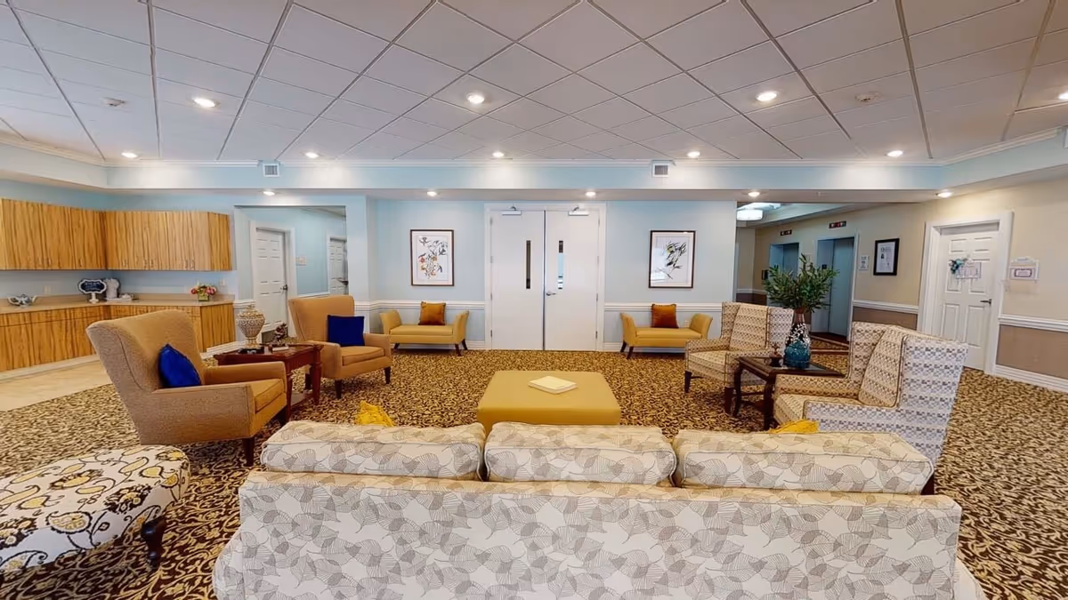 A spacious and well-lit common area in a senior living facility featuring multiple upholstered chairs and sofas arranged around a central ottoman. The room has patterned carpet, light blue walls with white trim, framed artwork, and a ceiling with recessed lighting. There are two white double doors in the center background, a kitchenette area with wooden cabinets on the left, and two elevators on the right side.
