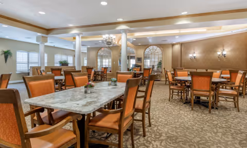 Large elegant dining room with marble-topped tables, upholstered chairs, chandeliers, columns and patterned carpet.