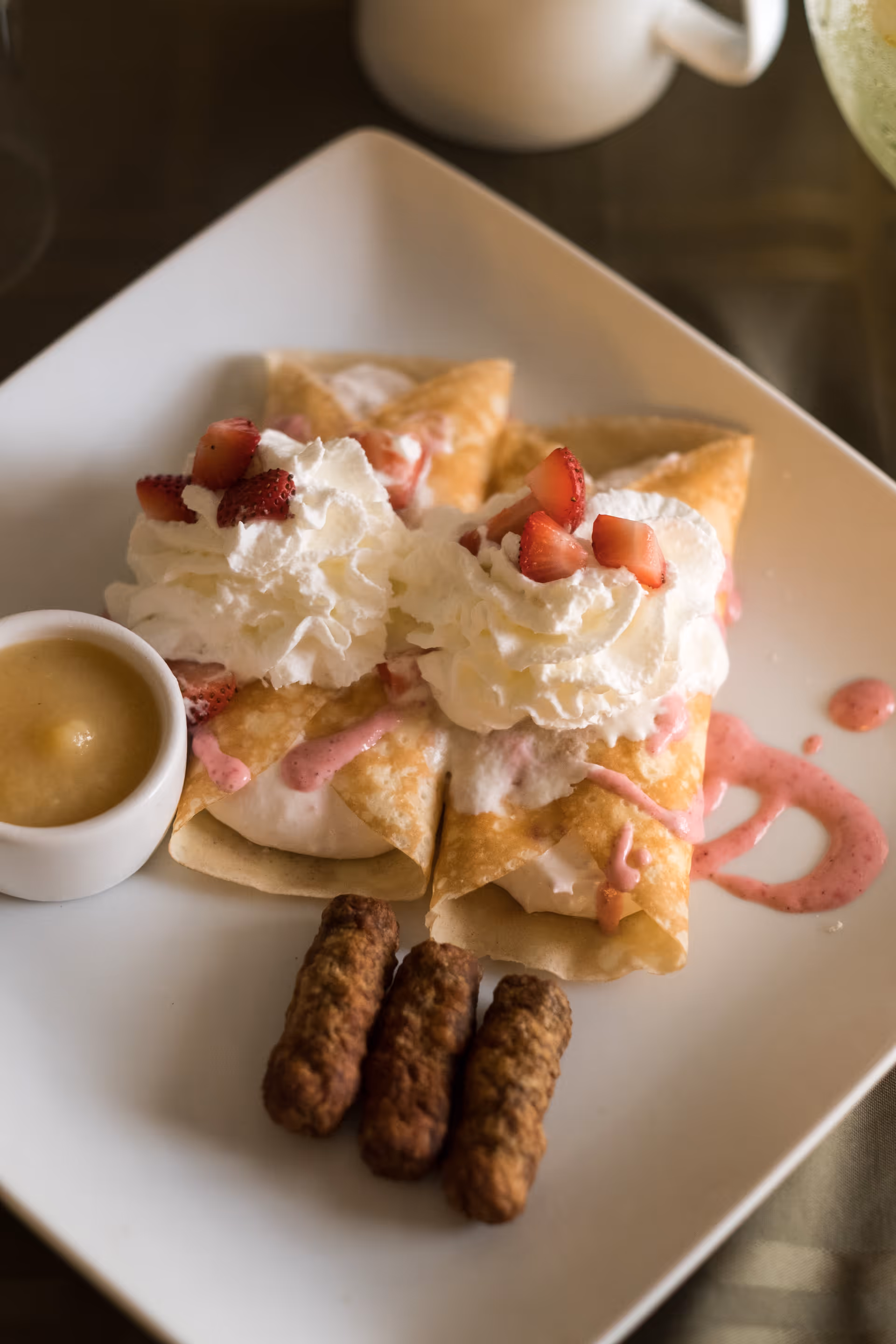 A white square plate with two crepes topped with whipped cream and chopped strawberries, drizzled with pink sauce. Three sausage links and a small cup of applesauce are also on the plate. A white mug is partially visible in the background.