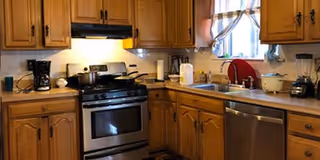 A kitchen with wooden cabinets, a stainless steel gas stove with a pot on it, a coffee maker, a blender, a dishwasher, and a window with a curtain above the sink.
