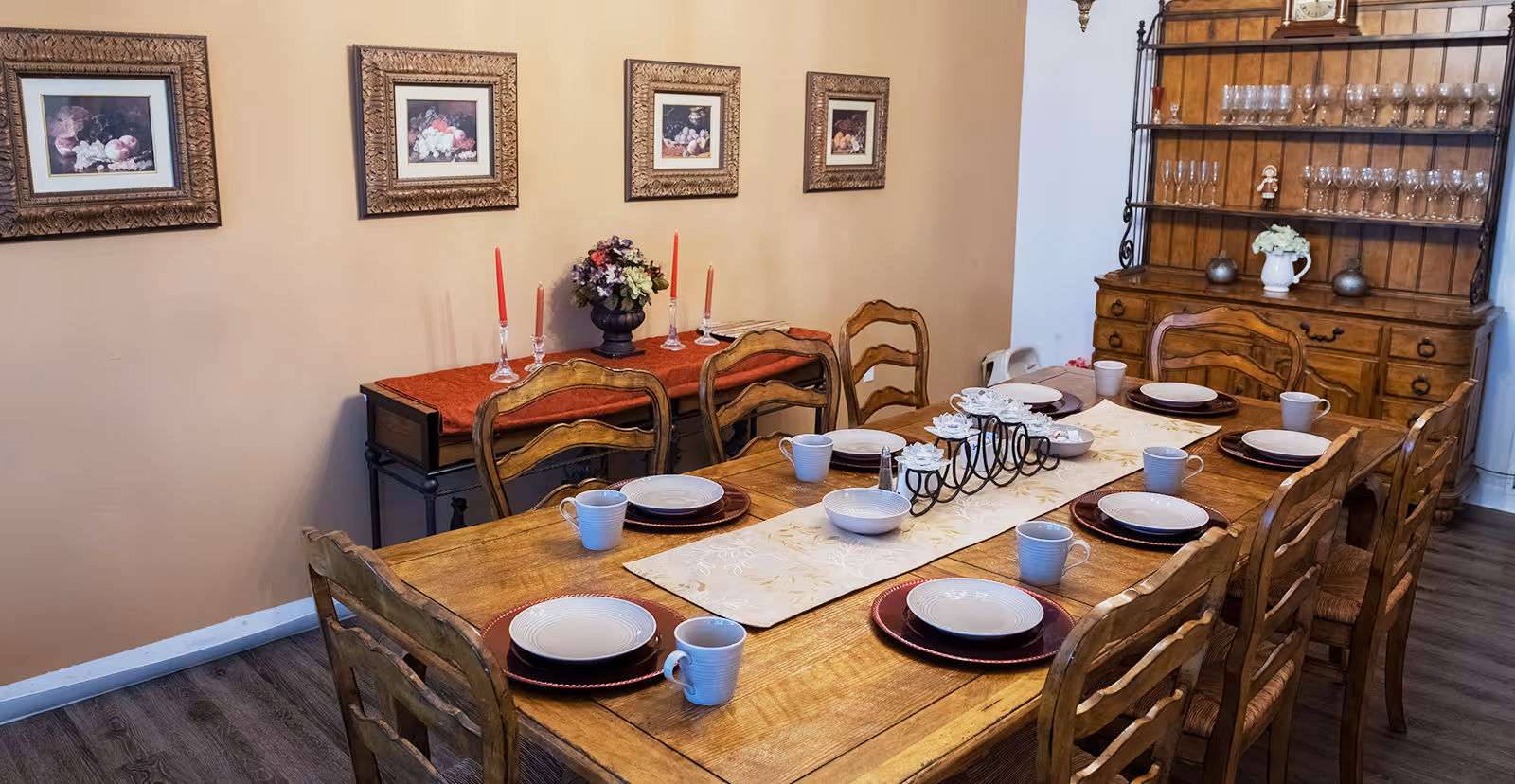 A dining room with a wooden table set for eight people with plates, bowls, and mugs. The table has a cream-colored runner with a decorative candle holder centerpiece. Behind the table, there is a wooden hutch filled with glassware and a sideboard with a red runner, candles, and a floral arrangement. Four framed paintings hang on the beige wall above the sideboard.