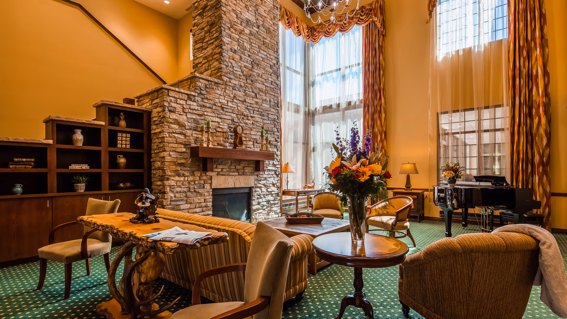A cozy living room with a tall stone fireplace, large windows with patterned curtains, a grand piano, and comfortable seating including armchairs and a sofa. A wooden table with a vase of flowers is in the foreground, and shelves with decorative items are visible on the left.