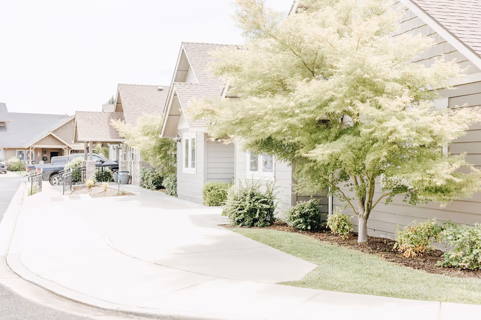 Exterior view of a senior living facility with multiple small buildings featuring peaked roofs, beige siding, and landscaped greenery including trees and shrubs along a paved walkway and driveway.
