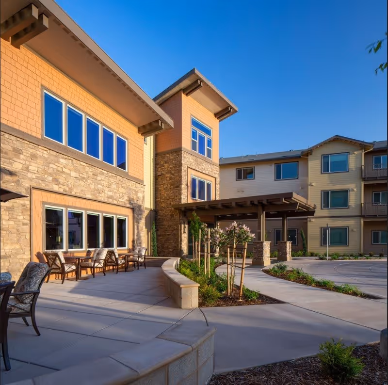 Outdoor patio area of a senior living facility with stone and wood exterior walls, several chairs arranged along the patio, small trees and plants in landscaped beds, and a covered entrance under a clear blue sky.