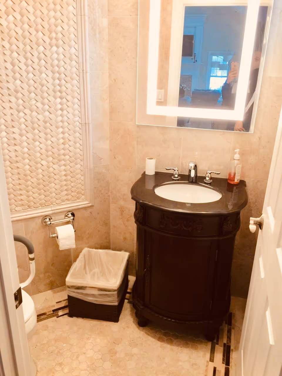 A small bathroom with beige tiled walls and floor. There is a dark wooden vanity with a black countertop and a white sink. Above the sink is a rectangular mirror with built-in lighting. A soap dispenser and a roll of toilet paper are on the countertop. To the left, there is a toilet with a grab bar and a toilet paper holder mounted on the wall. Below the toilet paper holder is a trash bin with a plastic liner. The bathroom door is partially open, and a reflection of a person taking the photo is visible in the mirror.