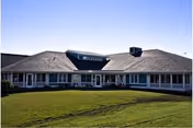 Single-story building with a wide, sloped roof and multiple windows, surrounded by a large grassy lawn under a clear blue sky.