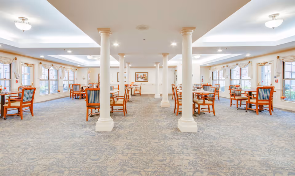 A spacious dining room with multiple wooden tables and chairs arranged neatly. The room features large windows with draped curtains allowing natural light to fill the space. White columns run down the center of the room, and the ceiling has recessed lighting and decorative fixtures. The carpet has a subtle patterned design in shades of blue and gray.