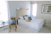 A bright bedroom with a neatly made bed featuring white linens and decorative pillows. There is a wooden chair with a patterned cushion next to the bed, a small table with books and a cup, two wall-mounted lamps above the headboard, and a framed picture on the wall. Light streams in through two windows with white curtains.