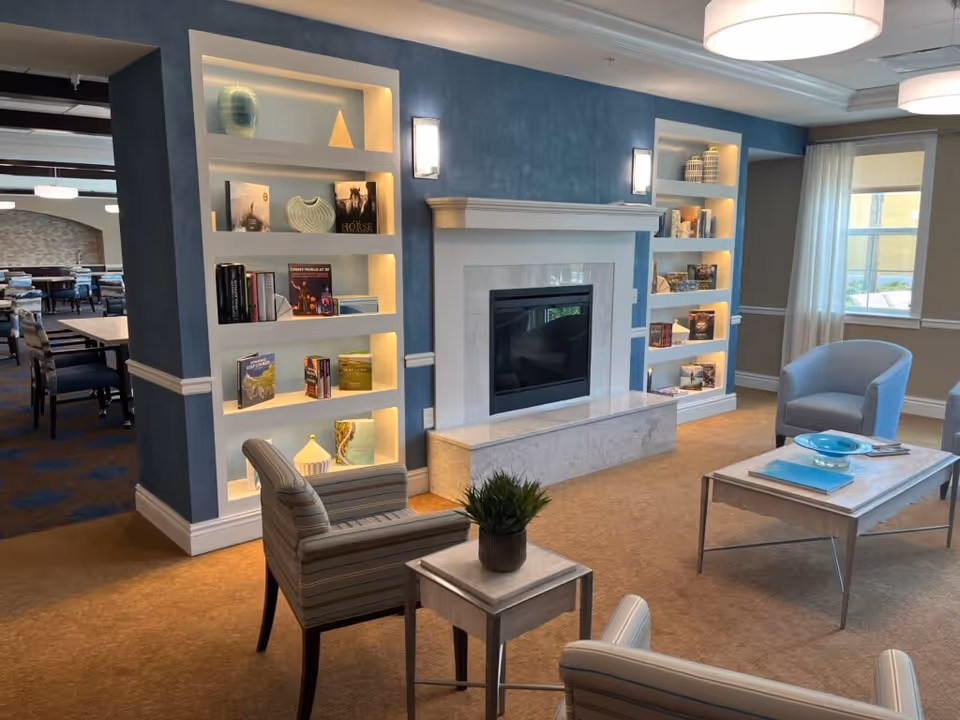 A cozy living room area in a senior living facility featuring a fireplace with a marble surround, built-in illuminated shelves displaying books and decorative items, two striped armchairs, a small table with a plant, a coffee table with a blue bowl and books, and a blue armchair near a window with white curtains.