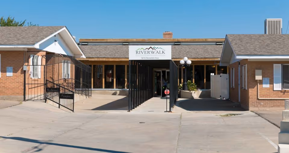 Front exterior view of Riverwalk Post Acute and Rehabilitation facility showing a single-story brick building with a covered entrance and a sign above the entrance displaying the facility name and address.