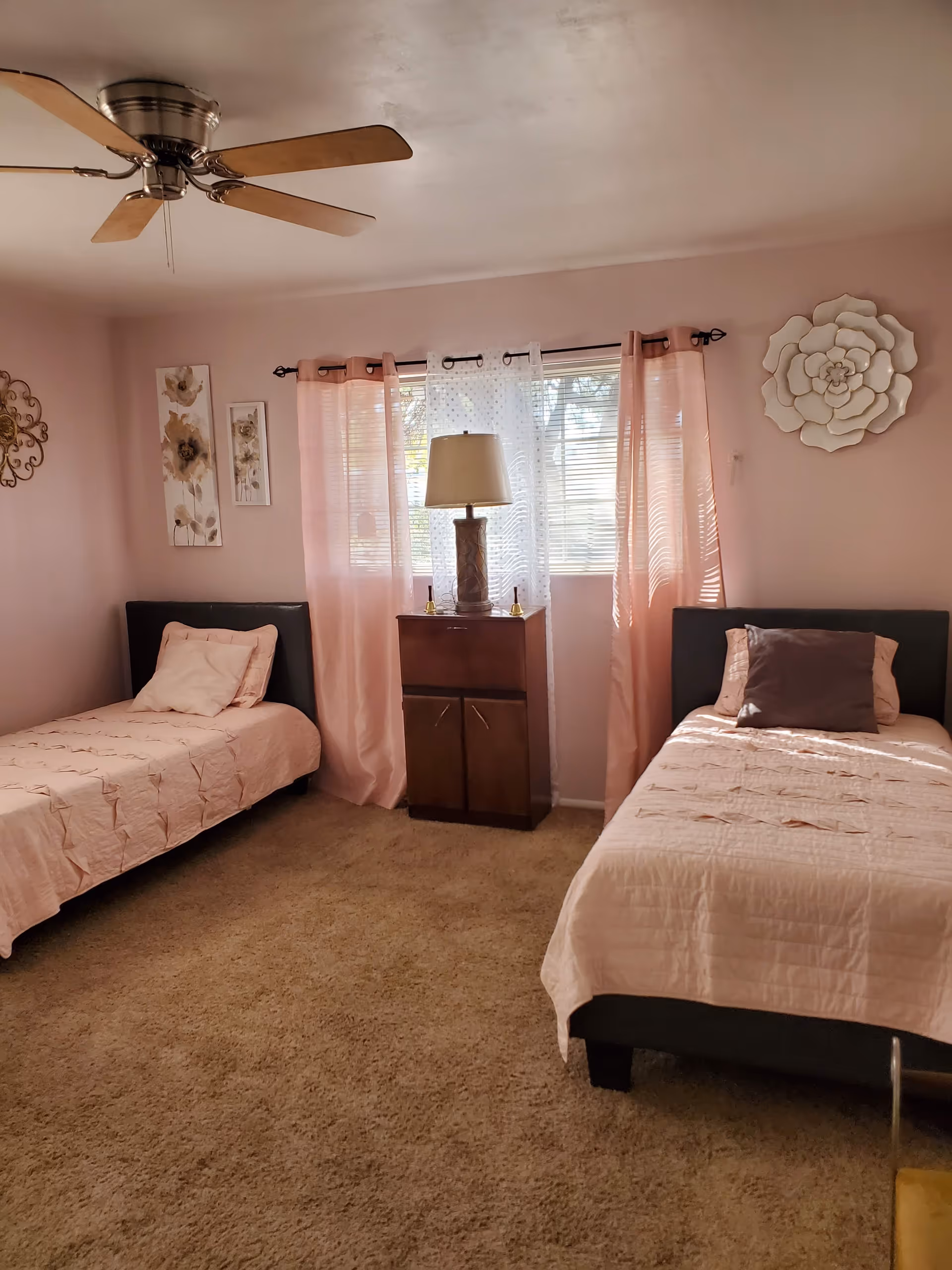 A bedroom with two single beds on either side of a window. The beds have light pink bedding and pillows. Between the beds is a small wooden cabinet with a lamp on top. The window has pink curtains and white sheer curtains. The walls are light pink with floral wall decorations, and there is a ceiling fan with wooden blades.