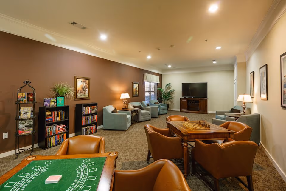 A spacious common lounge area with a card table and a chess table surrounded by brown leather chairs. The room has a brown accent wall with bookshelves filled with books and a plant. There are several light blue armchairs along the walls, a large flat-screen TV on a wooden stand, framed artwork on the walls, and table lamps providing warm lighting.