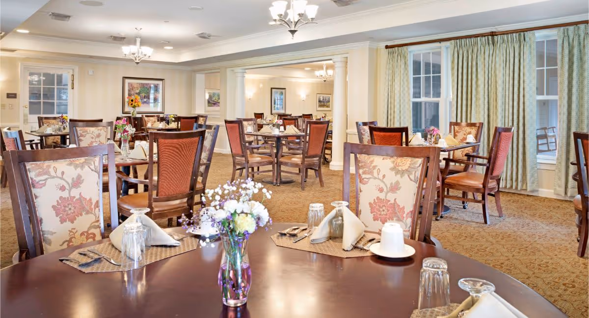 A bright and elegant dining room with multiple wooden tables and chairs. Each table is set with placemats, napkins, glasses, and silverware. There are floral centerpieces on the tables. The room has large windows with green curtains, beige walls, and carpeted floors. Ceiling lights and chandeliers provide warm lighting.