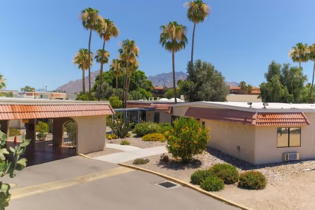 Exterior view of Casas Adobes Post Acute Rehabilitation Center showing single-story buildings with beige walls and red tile roofs, surrounded by desert landscaping with bushes and palm trees, with mountains visible in the background under a clear blue sky.