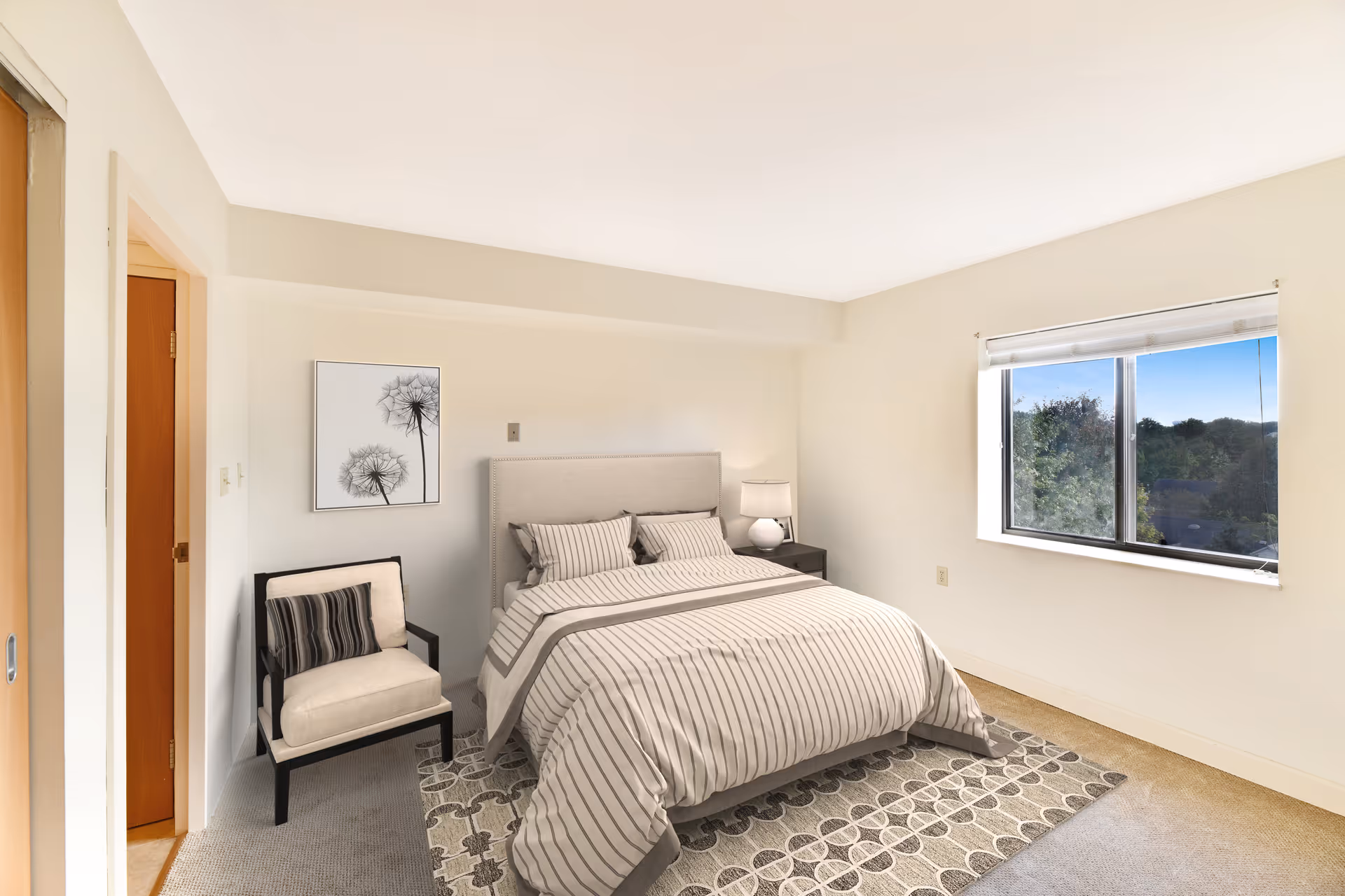 A bright bedroom with a large window showing a view of trees outside. The room features a bed with striped bedding, a nightstand with a lamp, a cushioned chair with a striped pillow, and a framed picture of dandelions on the wall. The walls are painted white and the floor is carpeted with a patterned rug under the bed.