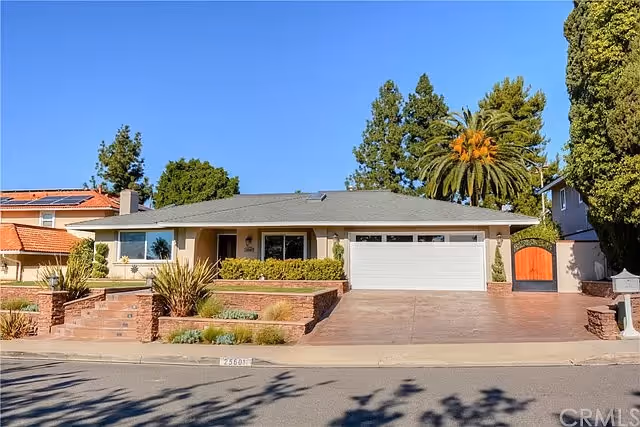 Single-story residential building with a gray roof, beige exterior walls, and a two-car garage. The front yard features brick planters with shrubs and plants, a concrete driveway, and a sidewalk. Tall trees and a clear blue sky are visible in the background.