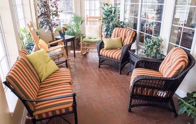 A bright sunroom with large windows featuring wicker furniture including a striped cushioned loveseat, two matching armchairs, and a wooden rocking chair with green and yellow accent pillows. The room has a terracotta tile floor and several potted plants adding greenery.