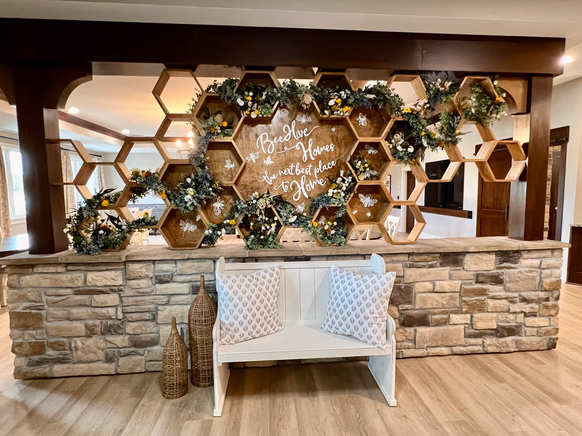Decorative honeycomb wooden divider with floral arrangements and a 'Beehive Homes' sign above a stone counter and a white bench with pillows in a common room.