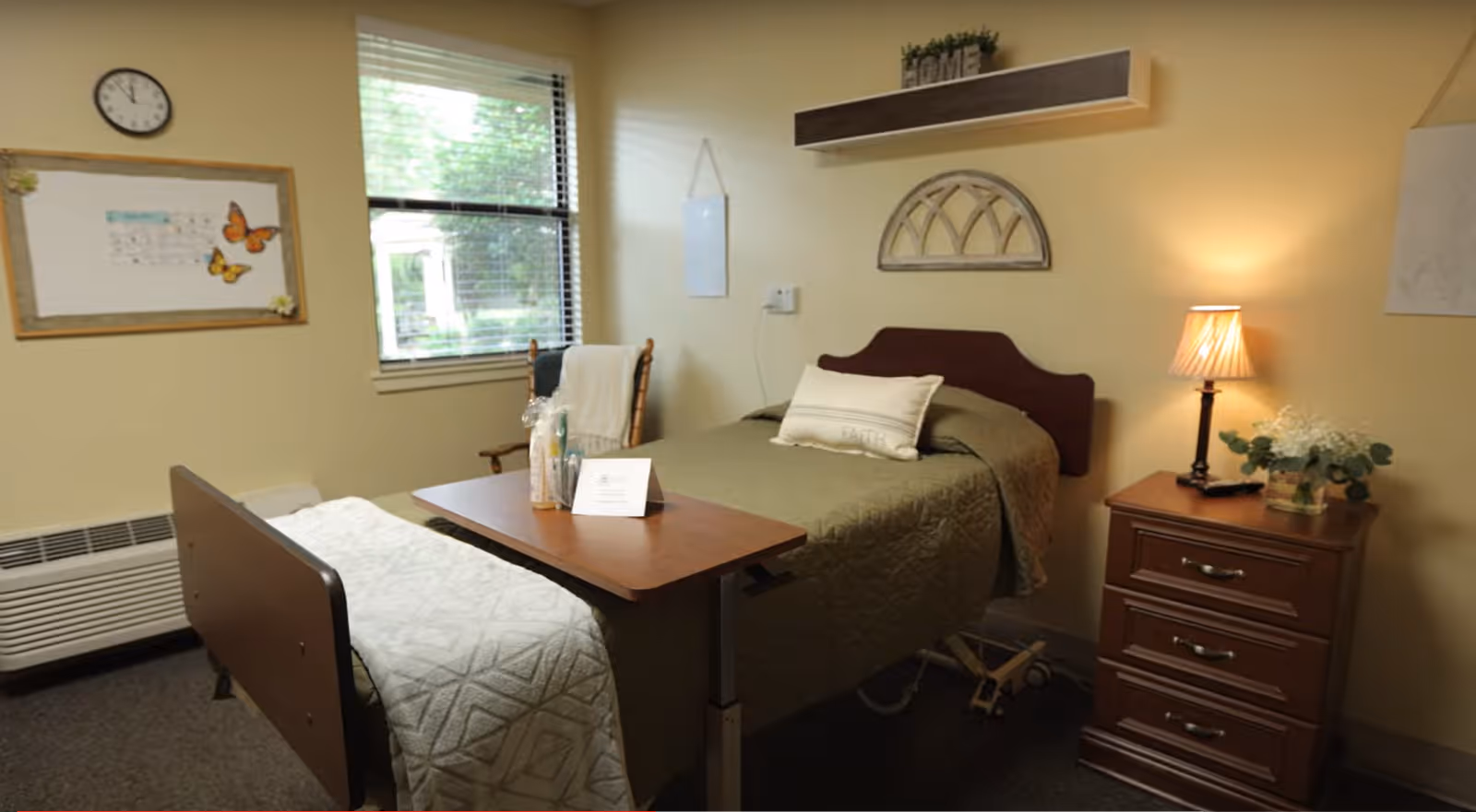 A small, cozy bedroom in a senior living facility with a single bed covered in a green quilt and a white pillow. There is a wooden bedside table with a lamp and a small plant. A wooden chair with a white blanket is placed near a window with blinds. A wall clock and a bulletin board with butterfly decorations hang on the wall. The room has soft yellow walls and a carpeted floor.