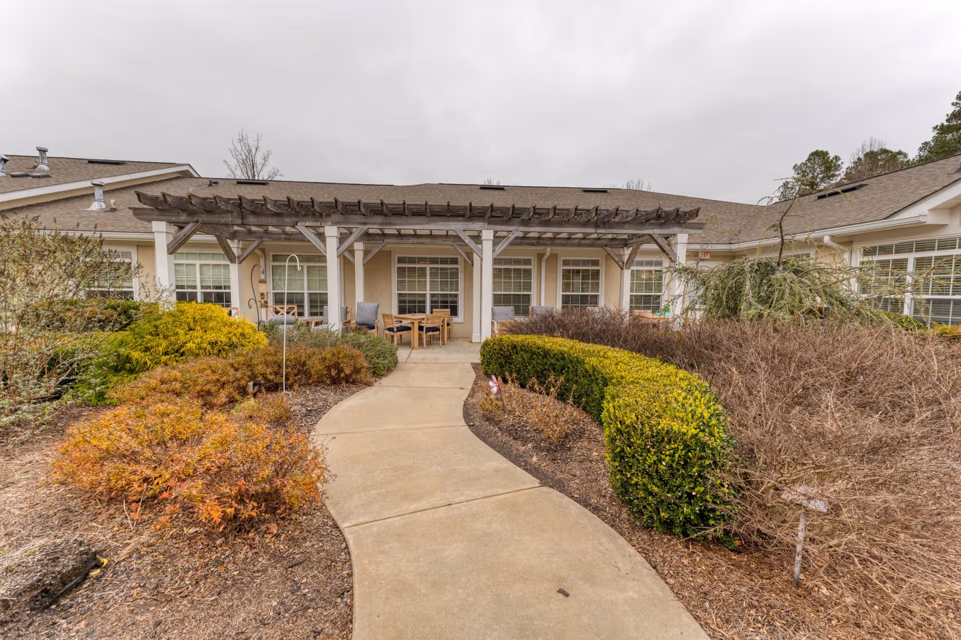 Outdoor garden area with a curved concrete pathway leading to a covered patio with wooden pergola. The patio has seating arrangements with chairs and a table. Surrounding the pathway are various bushes and shrubs, some green and some brown, under an overcast sky.