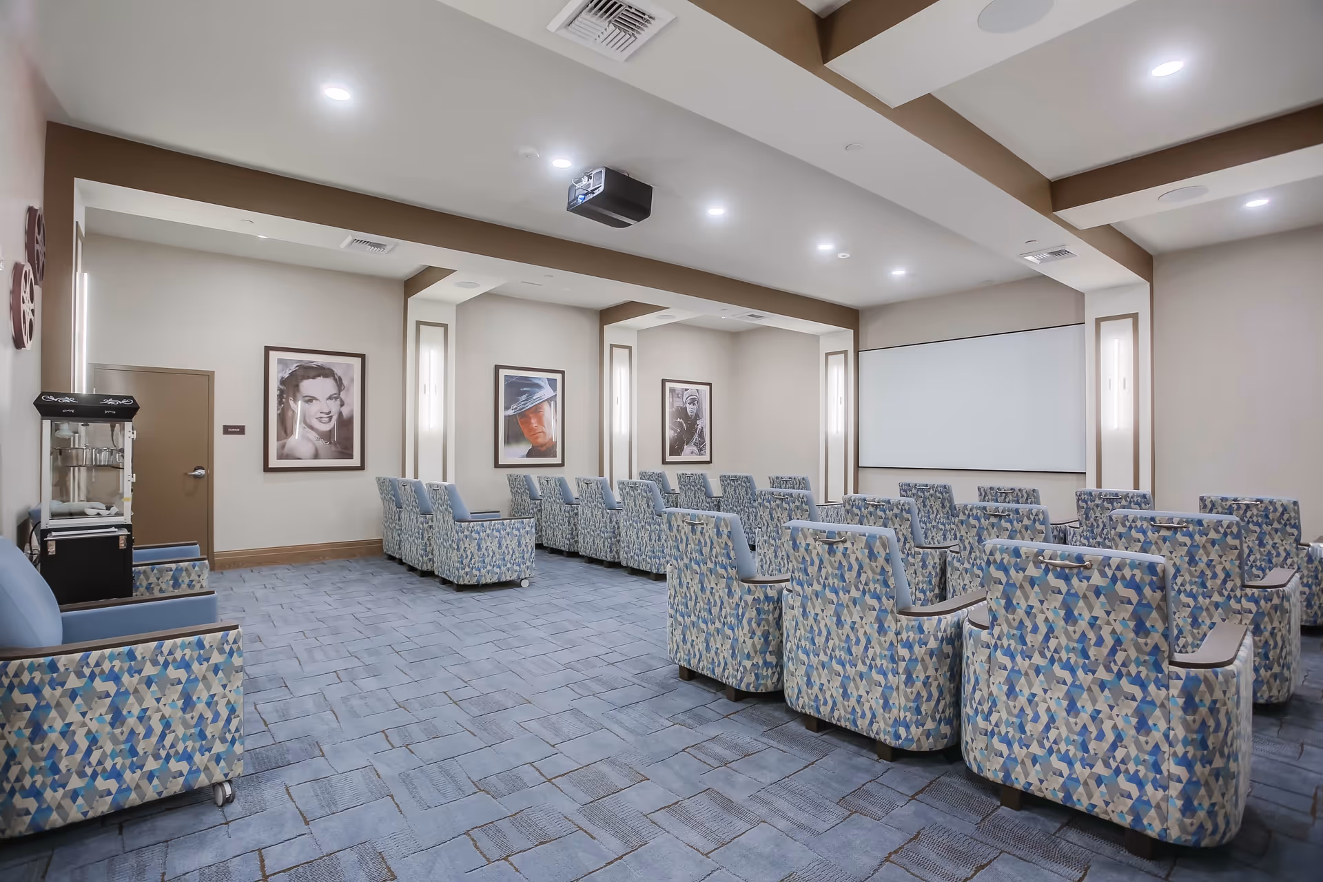 A small theater room with patterned blue and beige armchairs arranged in rows facing a large white projection screen. The room has beige walls with framed black-and-white and color portraits, recessed lighting in the ceiling, and a popcorn machine near the entrance door.