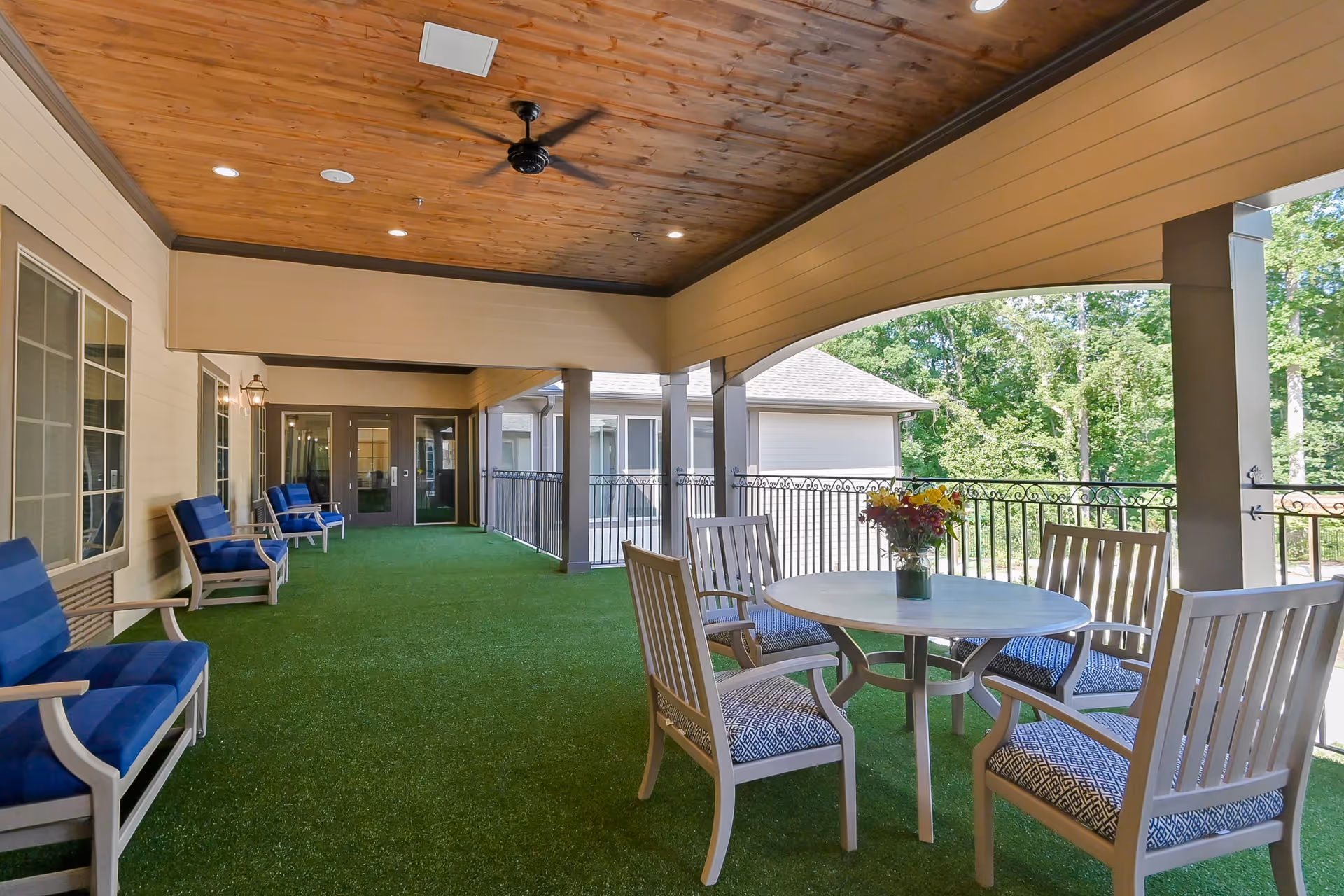 Covered outdoor seating area with cushioned chairs and a round table on green turf overlooking trees.