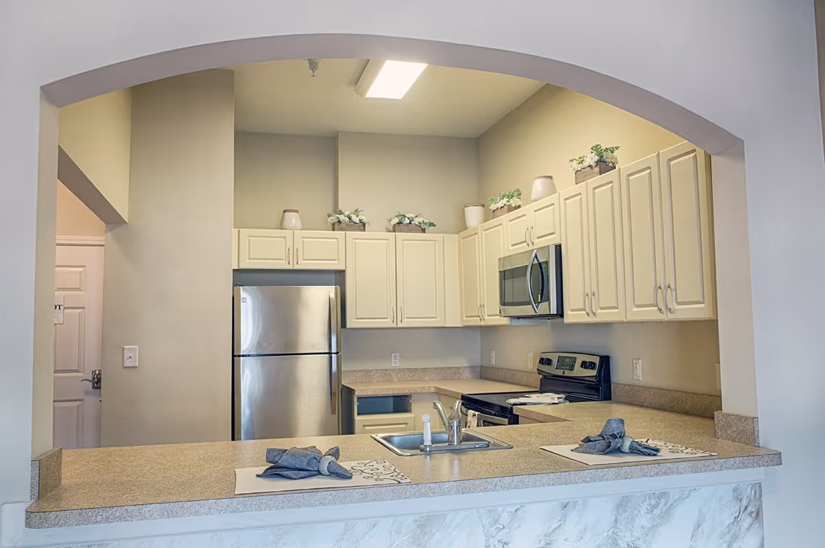 A modern kitchen with cream-colored cabinets, a stainless steel refrigerator, microwave, and stove. The kitchen has a beige countertop with a sink and two place settings with napkins. Decorative plants and vases are placed on top of the cabinets.