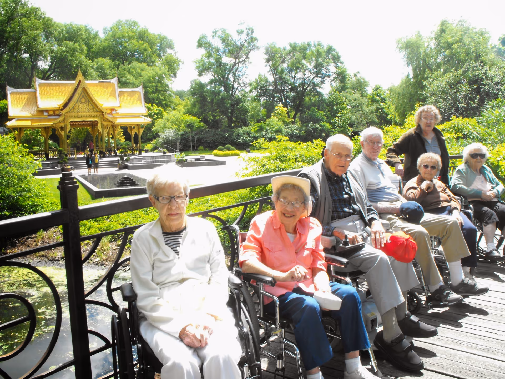 A group of elderly people, some in wheelchairs, sitting on a wooden deck outdoors near a pond with lush greenery and a golden pavilion in the background on a sunny day.