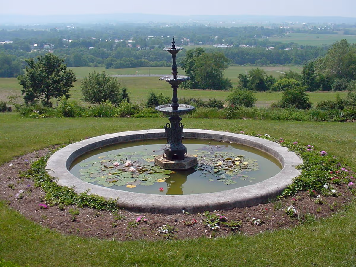 A circular stone fountain filled with lily pads sits on a grassy hill overlooking a rural landscape.