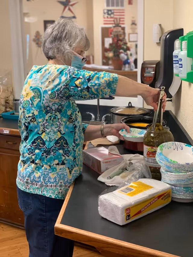 An elderly woman wearing a face mask and a colorful patterned shirt is serving food from a pot on a counter. The counter has disposable bowls, a jar of pickles, paper napkins, and a box of disposable gloves. The background shows a kitchen area with a sink and some decorations including an American flag and a star on the wall.