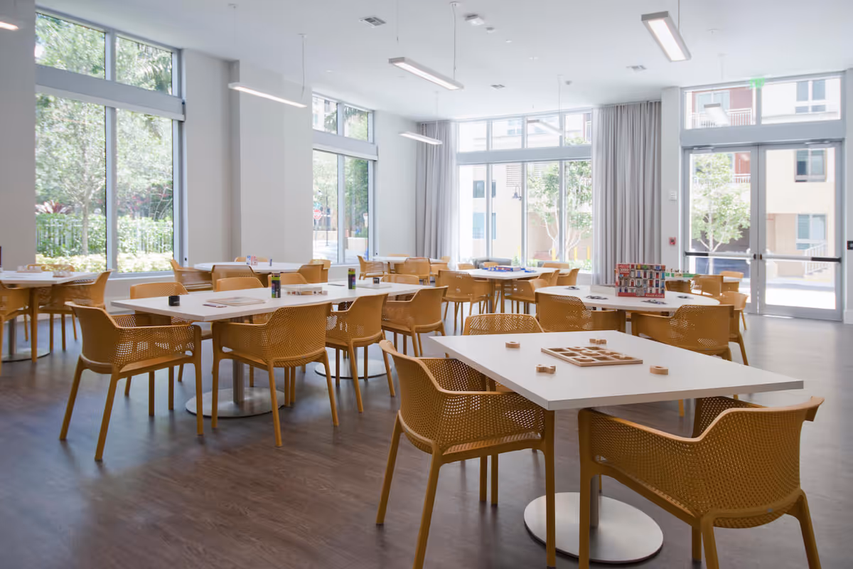 Bright communal dining/activity room with white tables, mustard-yellow chairs, and large windows letting in natural light.