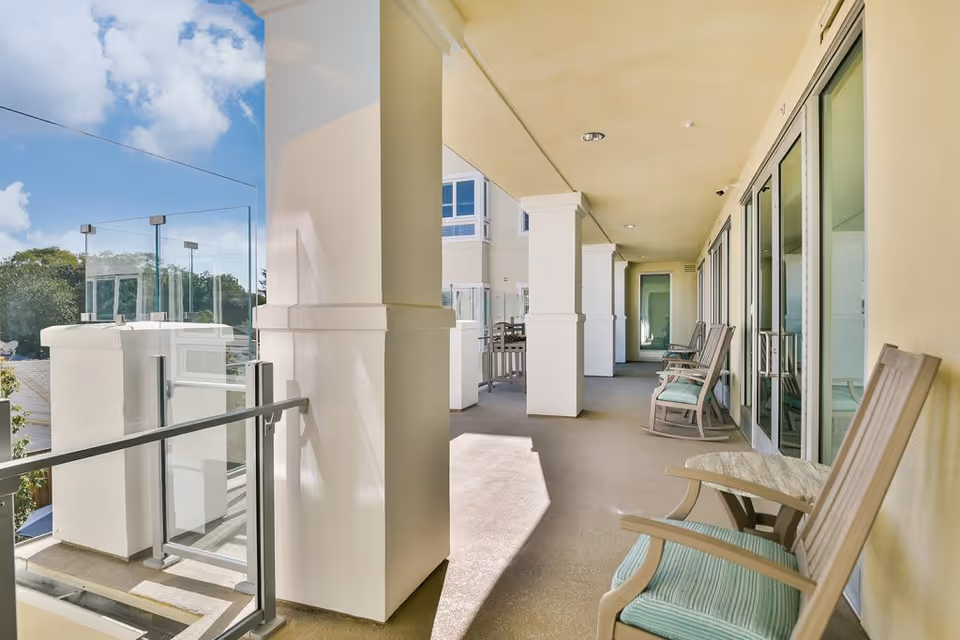 A covered outdoor balcony area with several cushioned wooden chairs and small tables along the right side, glass railings on the left side, and large white pillars supporting the ceiling. The sky is partly cloudy and visible beyond the balcony.